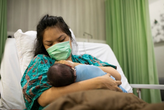 Mother with face-mask in hospital bed breastfeeding her baby.